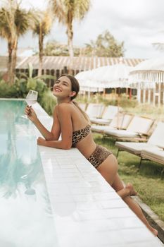 Smiling woman in leopard print swimwear enjoying a drink by a pool on a sunny day.