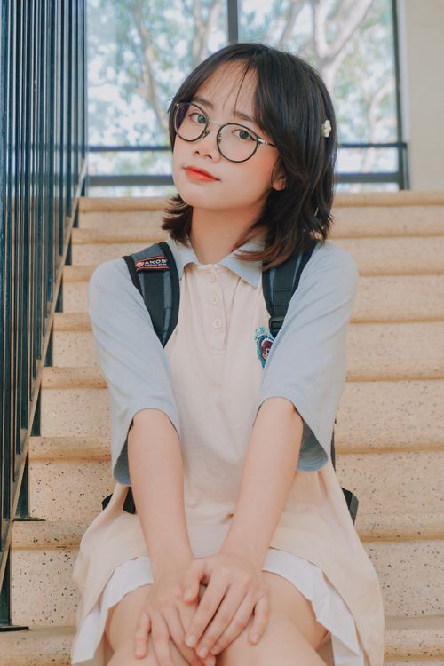 Free Charming portrait of a young female student sitting on a staircase, wearing eyeglasses and a uniform in Vietnam. Stock Photo