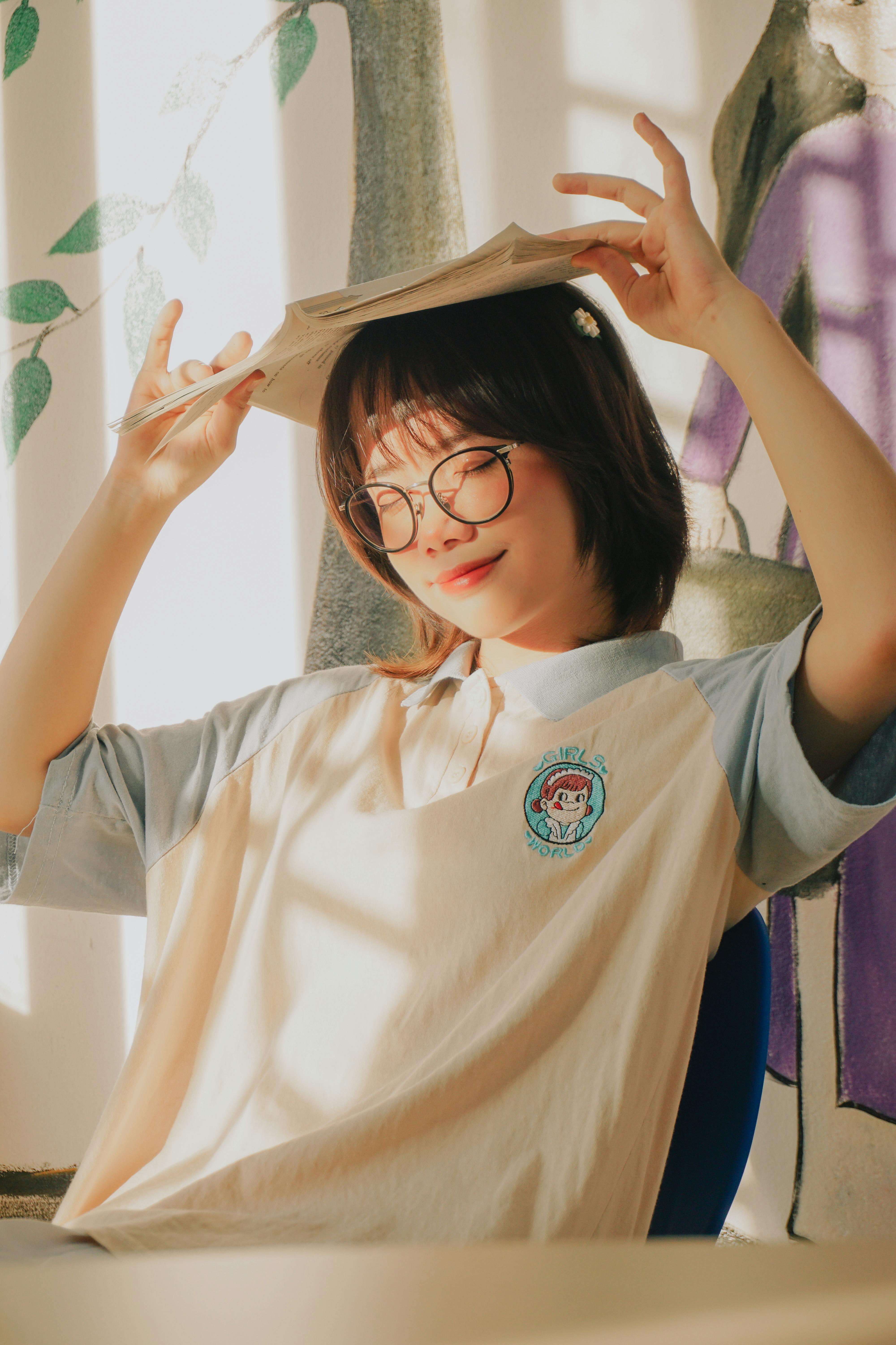 Portrait of a smiling Asian student with glasses and book on head, enjoying sunlight.