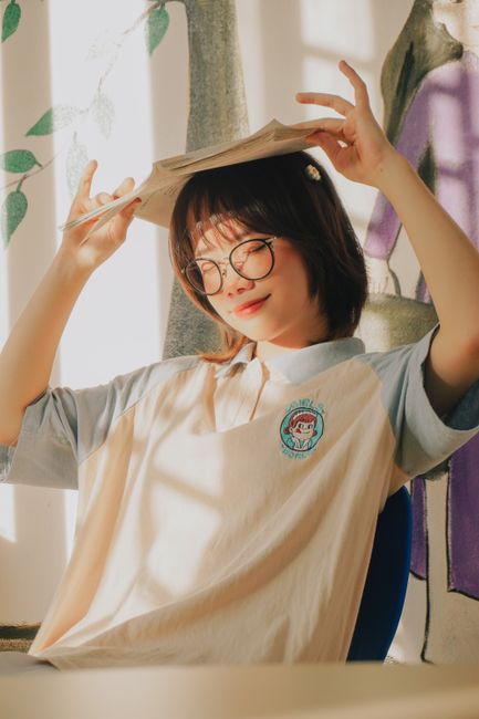 Portrait of a smiling Asian student with glasses and book on head, enjoying sunlight.