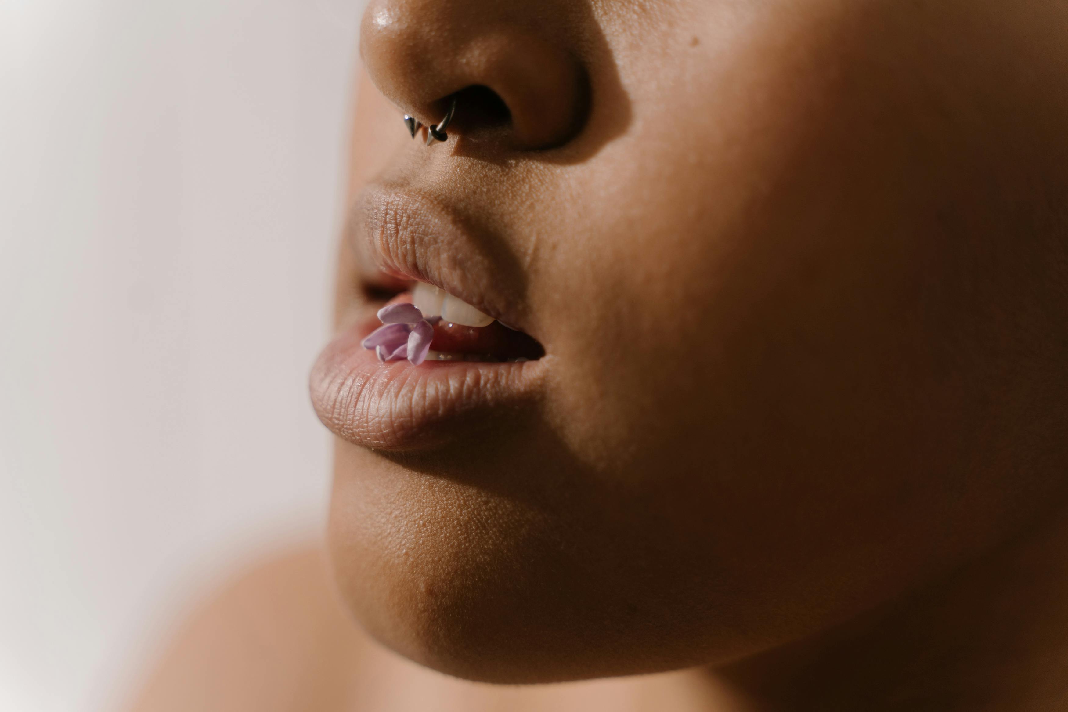Artistic close-up of lips with a nose ring and a purple flower held between the teeth.