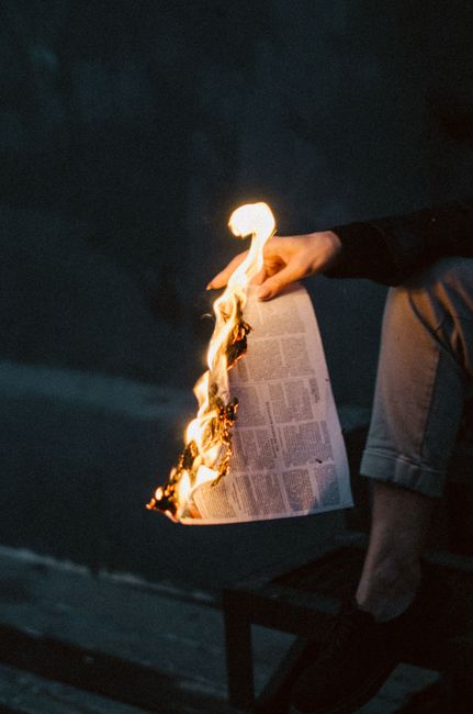 A close-up view of a newspaper set ablaze, held by an adult hand, showcasing fire's intensity at night.