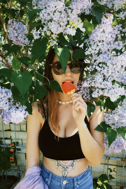 Young Woman Eating an Ice Lolly While Standing Under a Lilac Tree
