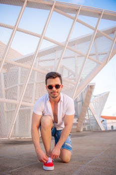 Caucasian man crouching outdoors, tying shoe against architectural backdrop wearing casual attire.
