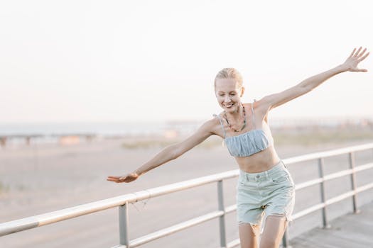 A young woman joyfully balances on railings by the beach during a sunny day.
