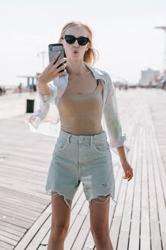 Blonde woman wearing sunglasses taking a selfie with her smartphone on a sunlit boardwalk.