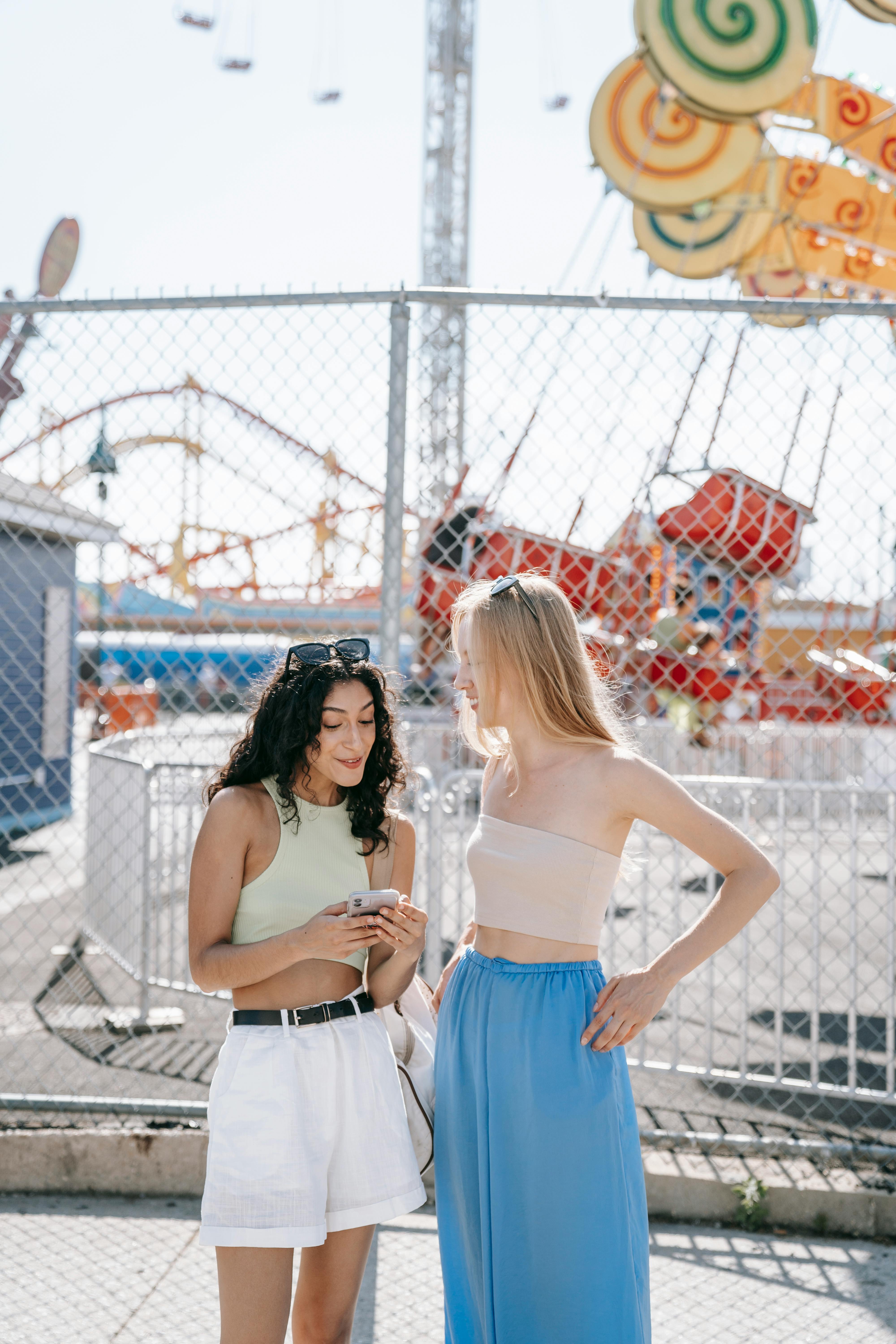 Two young women enjoy a sunny day at an amusement park, sharing a moment of fun and laughter.