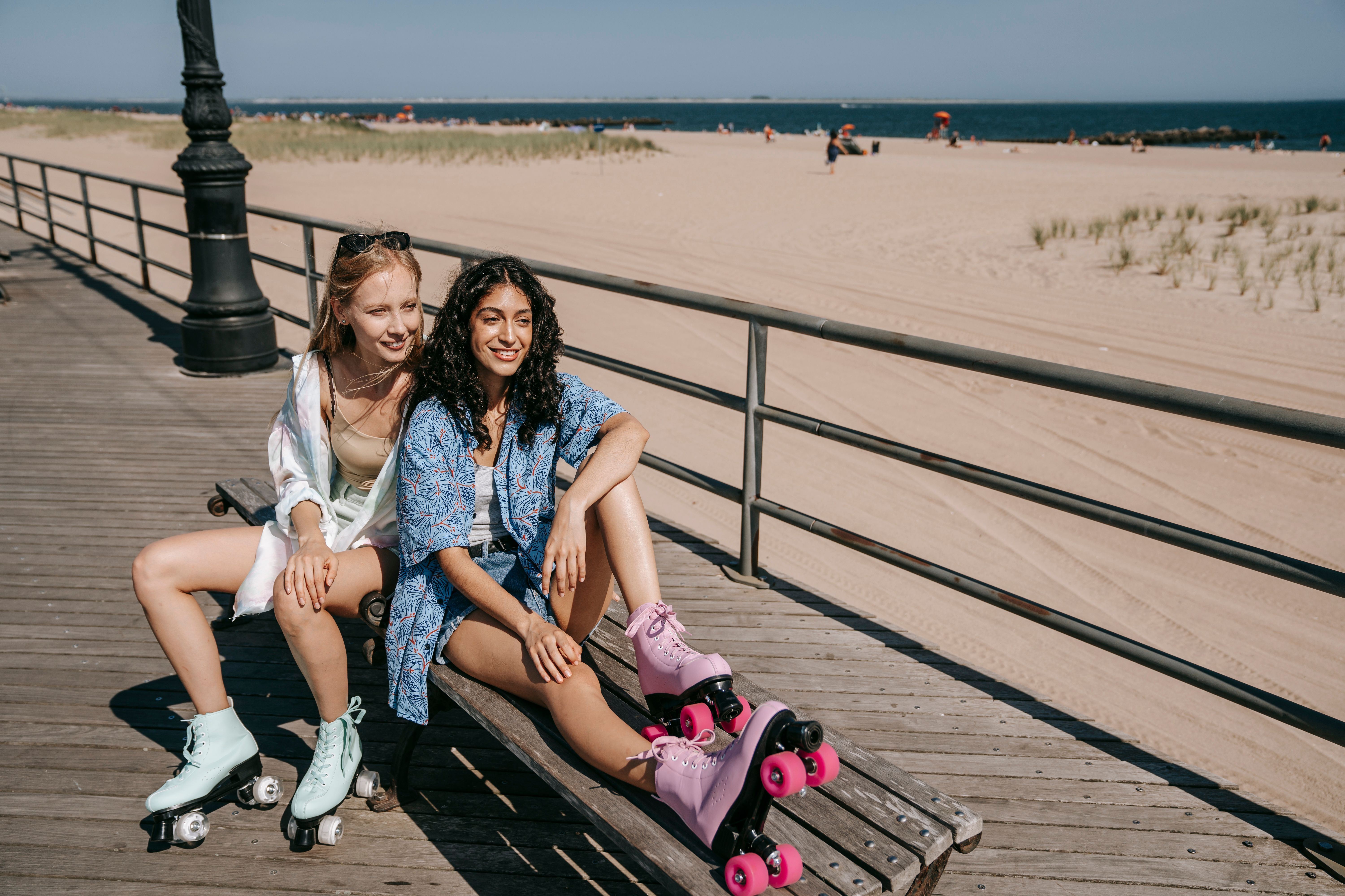 Two smiling women in roller skates enjoy a sunny beach day sitting on a bench.