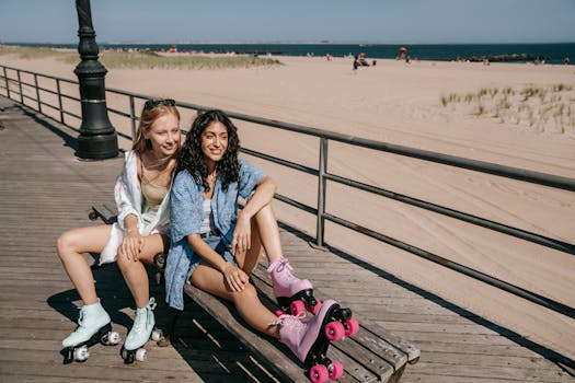 Two smiling women in roller skates enjoy a sunny beach day sitting on a bench.