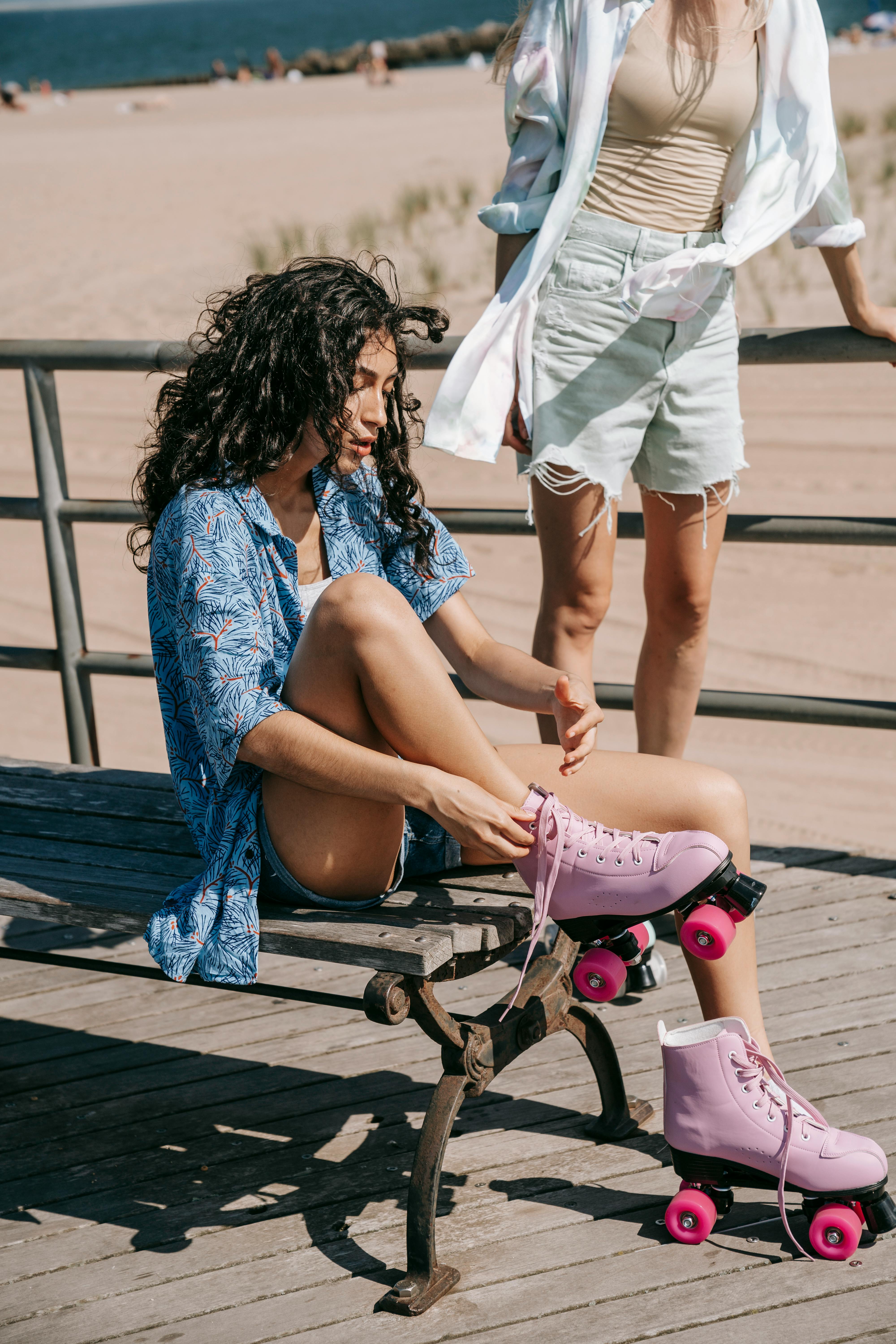 Two women, one sitting, putting on pink roller skates, beachside setting, outdoor fun.