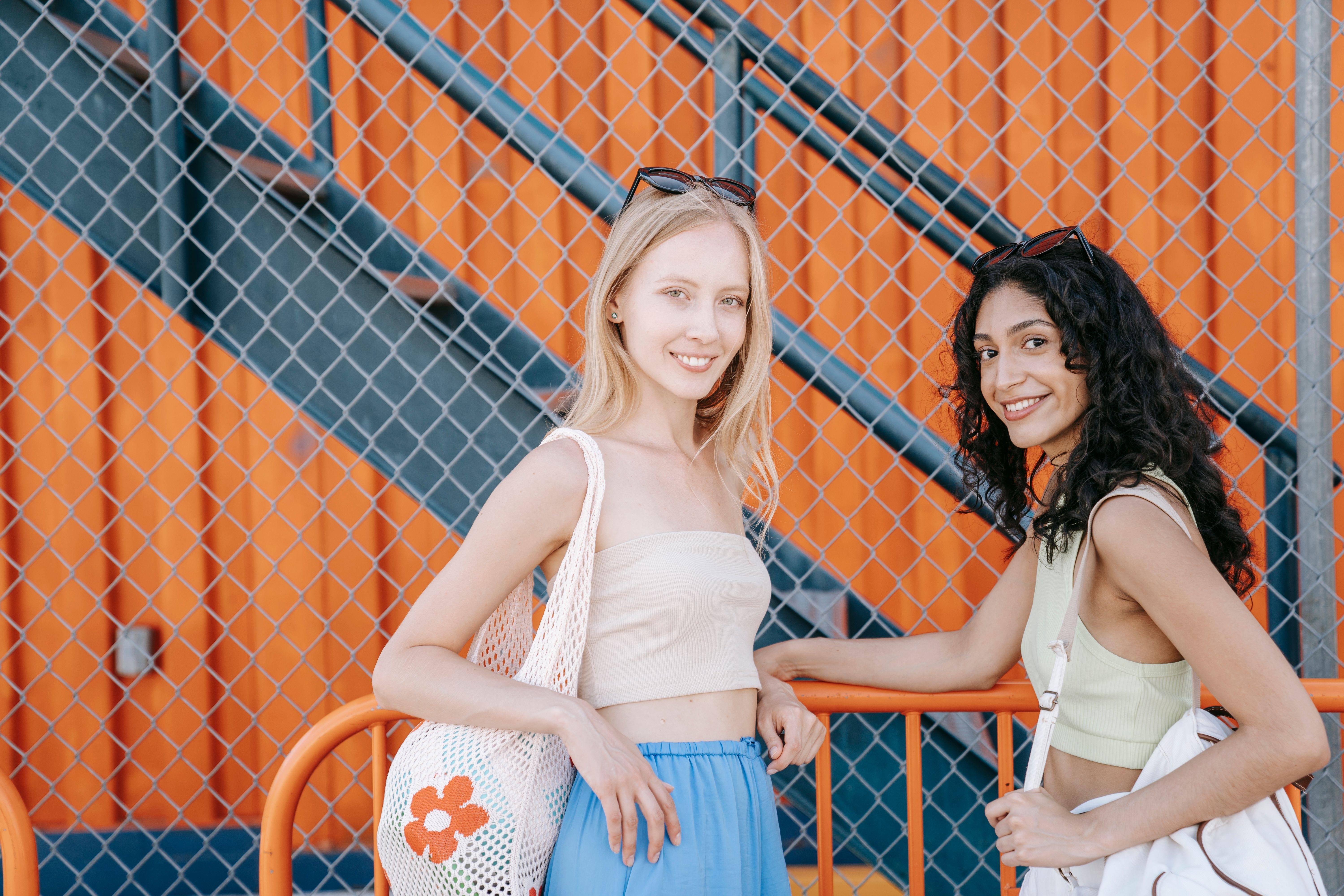 Two diverse young women smiling and posing by an orange chain link fence outdoors.