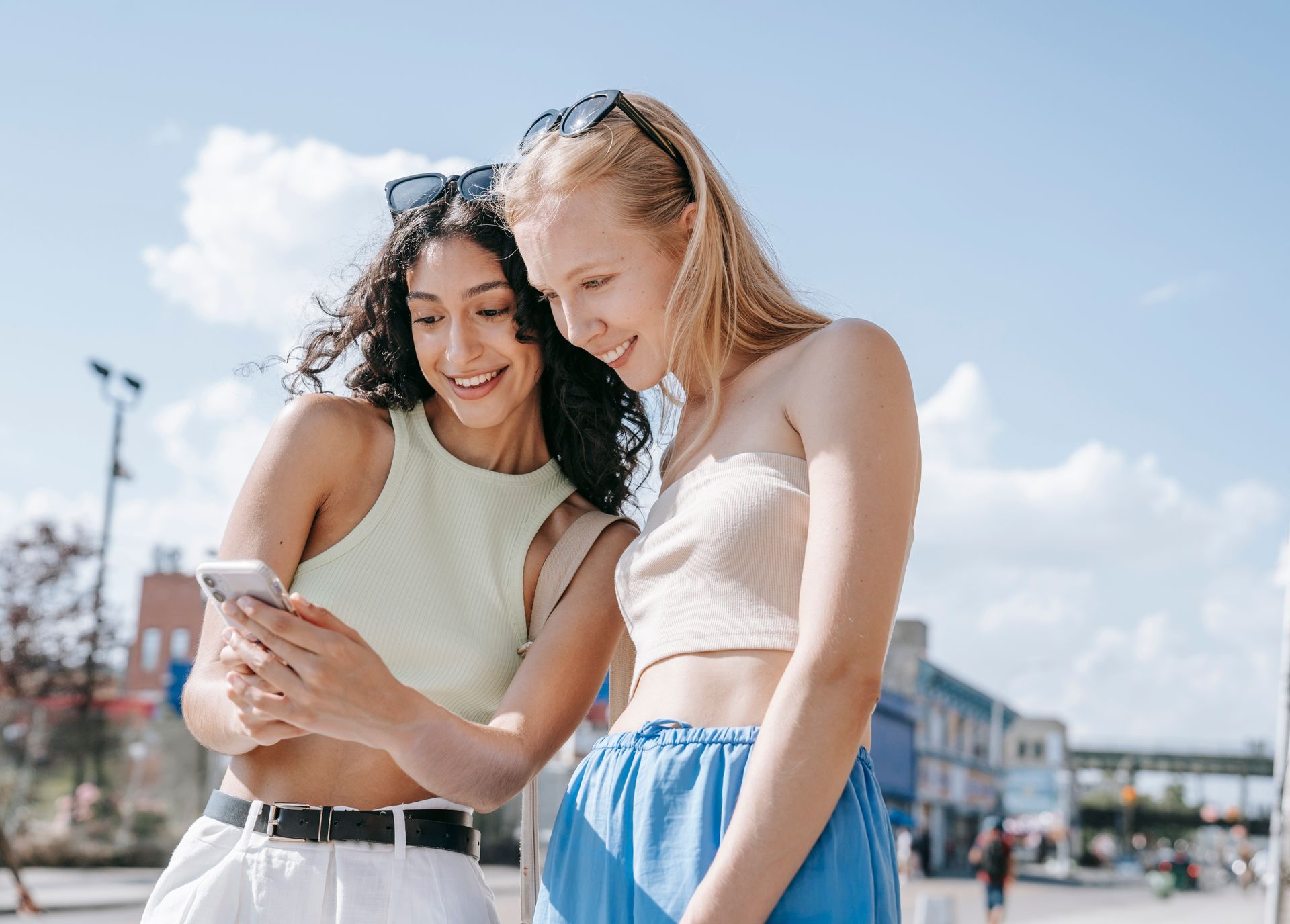 Two women smiling and checking a phone during a sunny day outdoors, enjoying their time together.