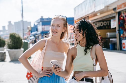 Free Two friends enjoying a sunny day outdoors, laughing and checking a smartphone on a bustling city street. Stock Photo