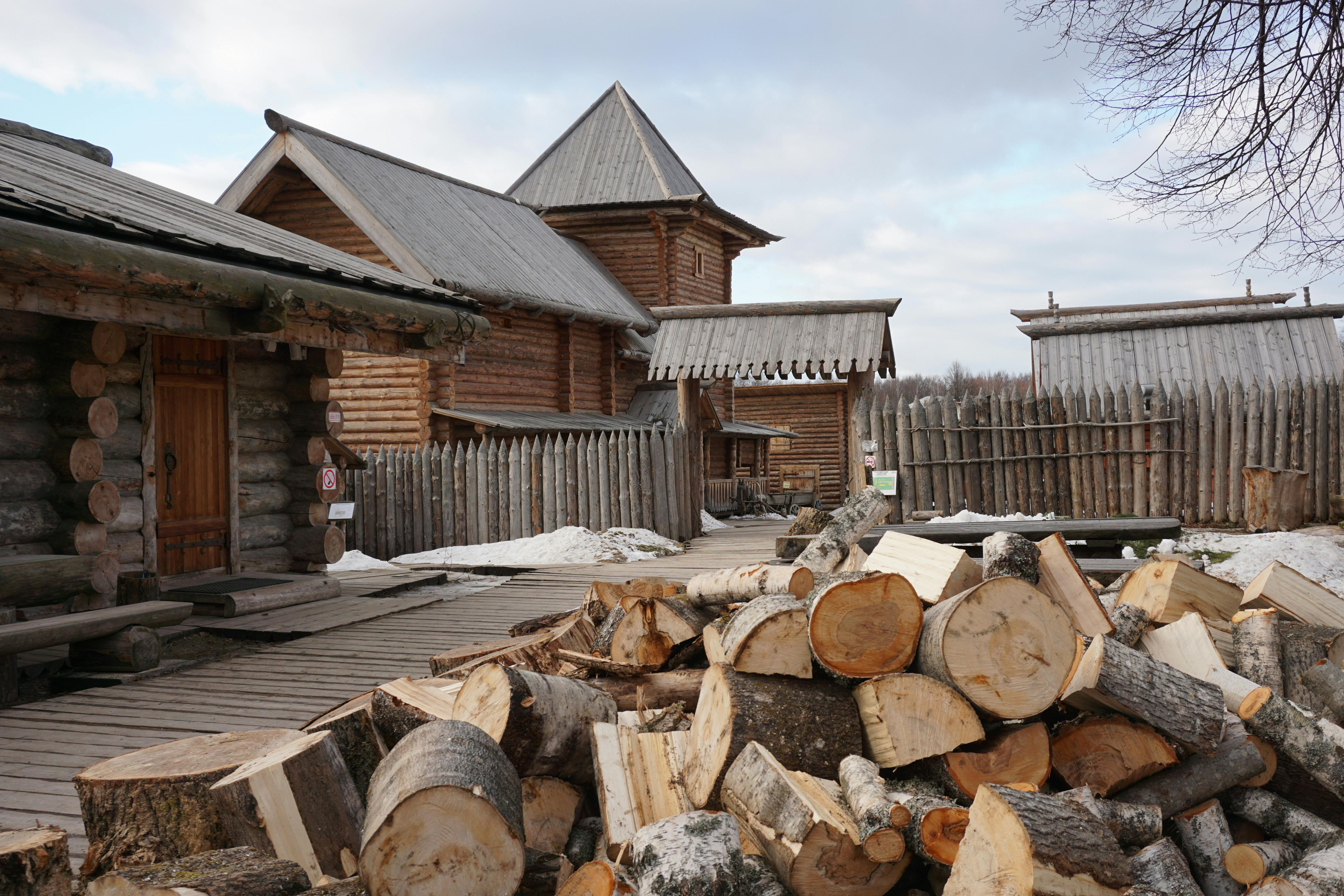 Chopped logs piled in front of traditional Russian wooden buildings, showcasing rustic architecture.