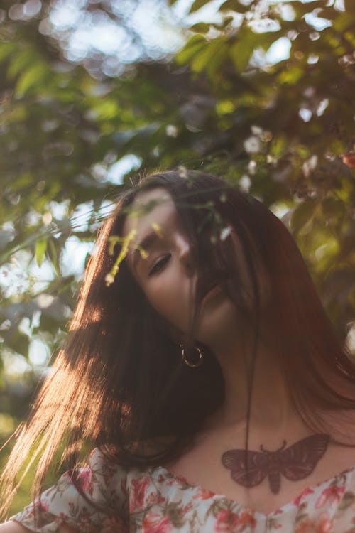 Young charming female with tattoo on chest with long dark hair dreaming among green leaves and branches of tree in sunlight
