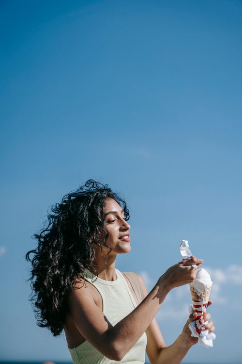 Free Young woman with curly hair enjoying a melting ice cream cone on a bright summer day by the shore. Stock Photo