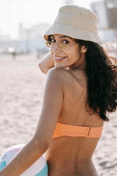 Young woman in bikini and bucket hat smiling at beach with colorful ball, enjoying sunny day.