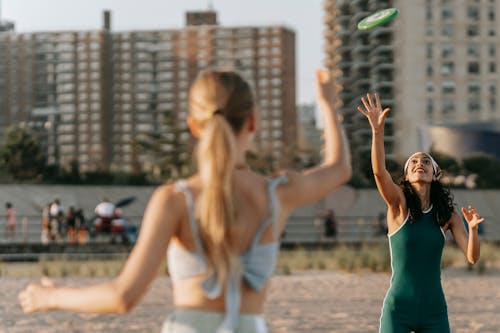 Free Two women enjoy a game of frisbee on a sunny beach day with a cityscape background. Stock Photo