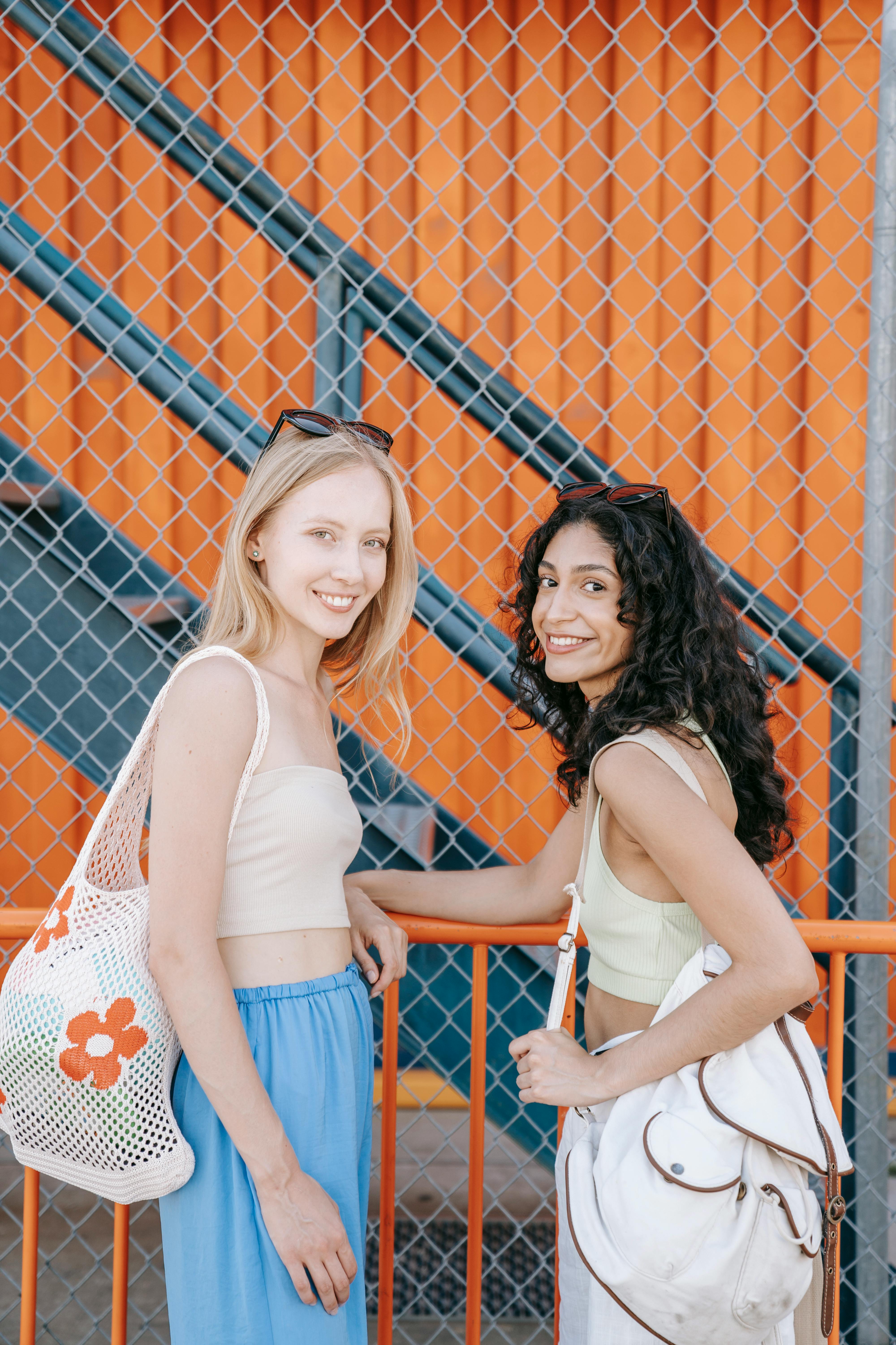 Two women smiling by a chain-link fence on a sunny day, embodying summer joy.