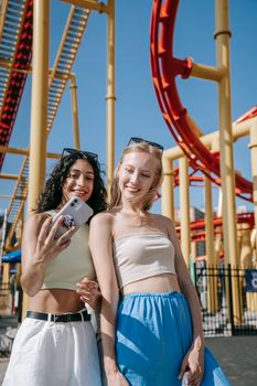Two young women taking a selfie in front of a roller coaster at an amusement park, capturing a joyful moment.