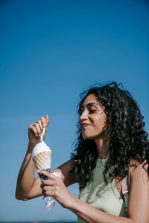 Free Smiling woman savoring ice cream cone under a clear blue sky during a joyful outdoor moment. Stock Photo