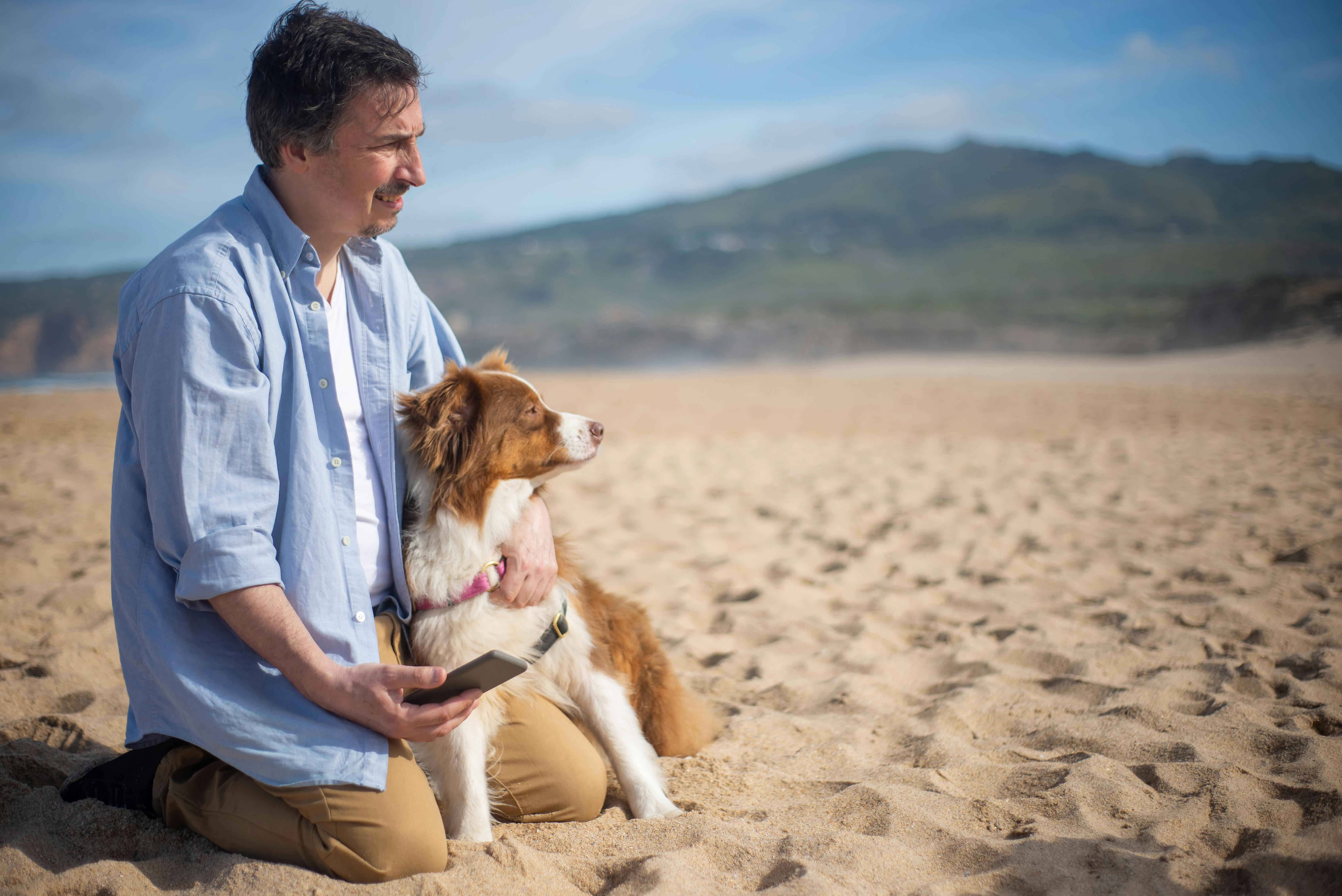 a man in blue polo shirt sitting on sand with a brown long coated dog