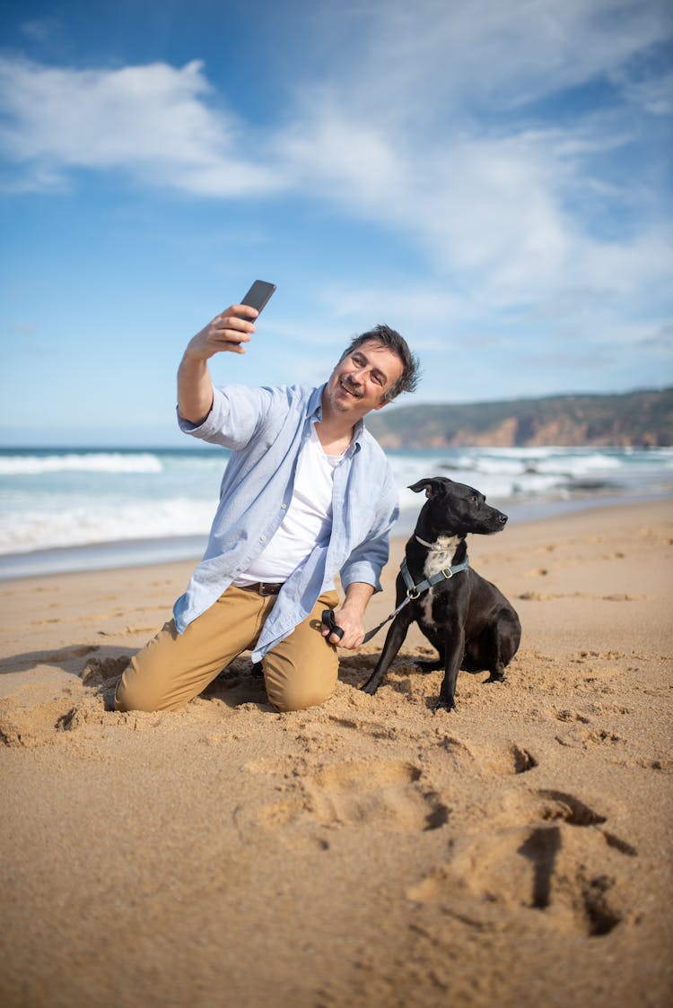 A Man In Blue Dress Shirt Sitting On Shore Taking A Selfie With A Black Short Coated Dog 