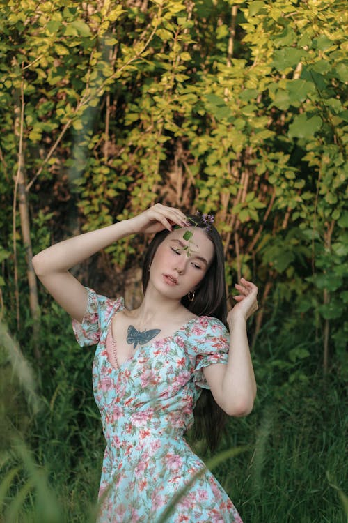 Young dreamy Asian female in silk floral dress touching face with sprig of meadow flowers while standing with raised hands in green bushes in summer garden