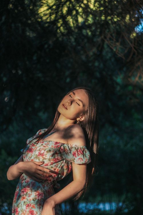 Free Young sensual female with long hair and wearing floral ornament dress with bare shoulders standing under tree in sunbeam Stock Photo