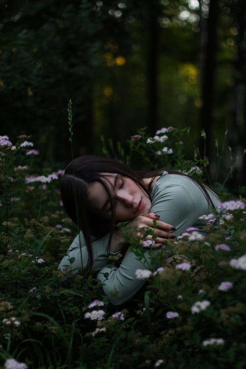 Free Young female with dark hair embracing herself and putting head on hands sitting on squat in grass with blooming meadow flowers in park Stock Photo