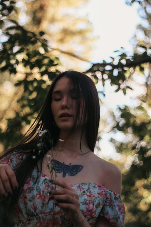 Young Asian woman with long dark hair and dress with bare shoulders with tattoo in chest standing with closed eyes and with field flowers in garden
