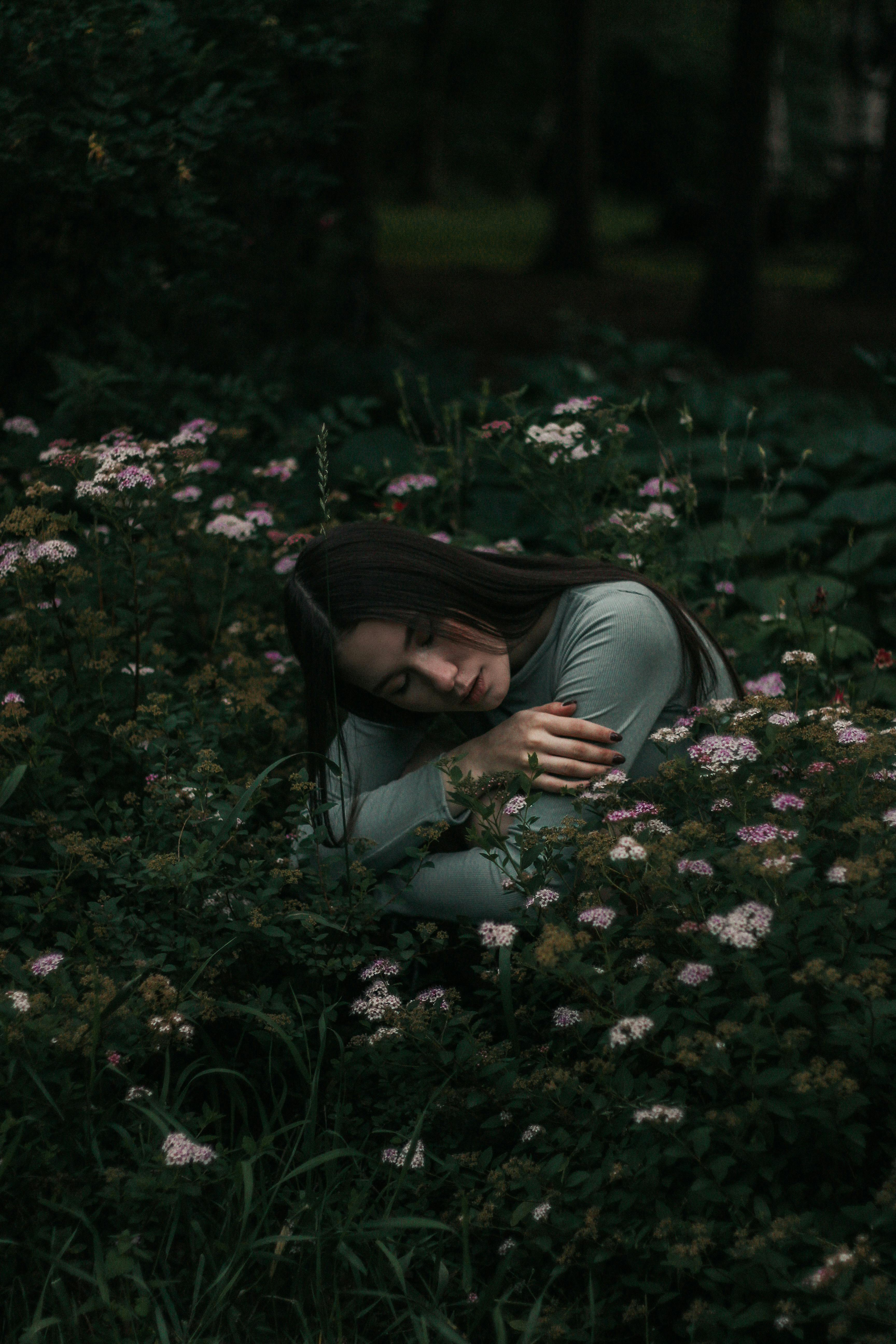 Young female with dark long hair embracing herself with closed eyes while sitting in blooming flowers in green forest