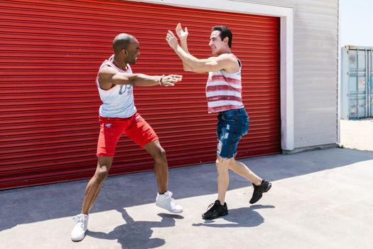Two men in casual attire high-fiving outside near a red roller shutter on a sunny day.