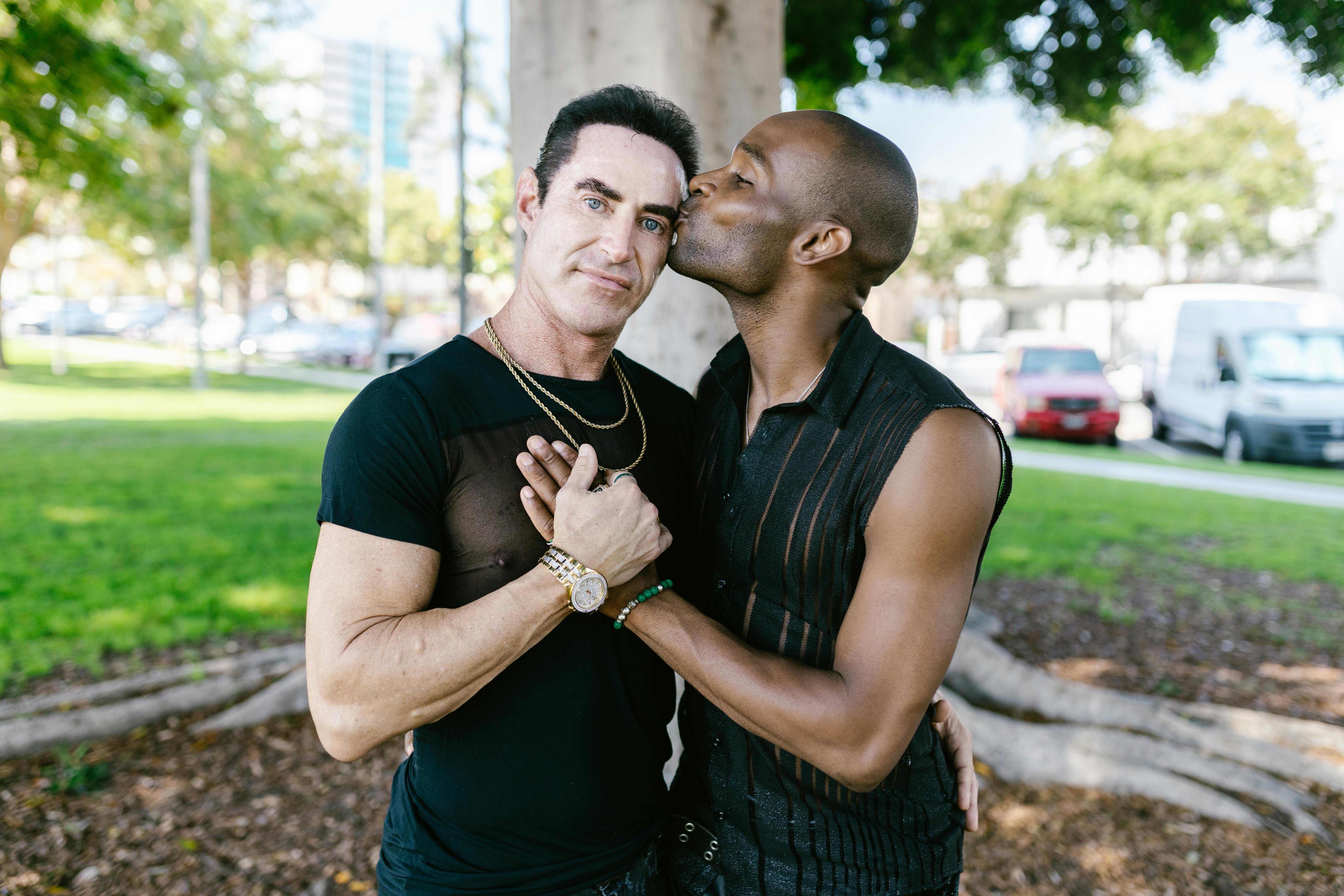 Affectionate interracial gay couple shares a tender moment in a sunny park setting.