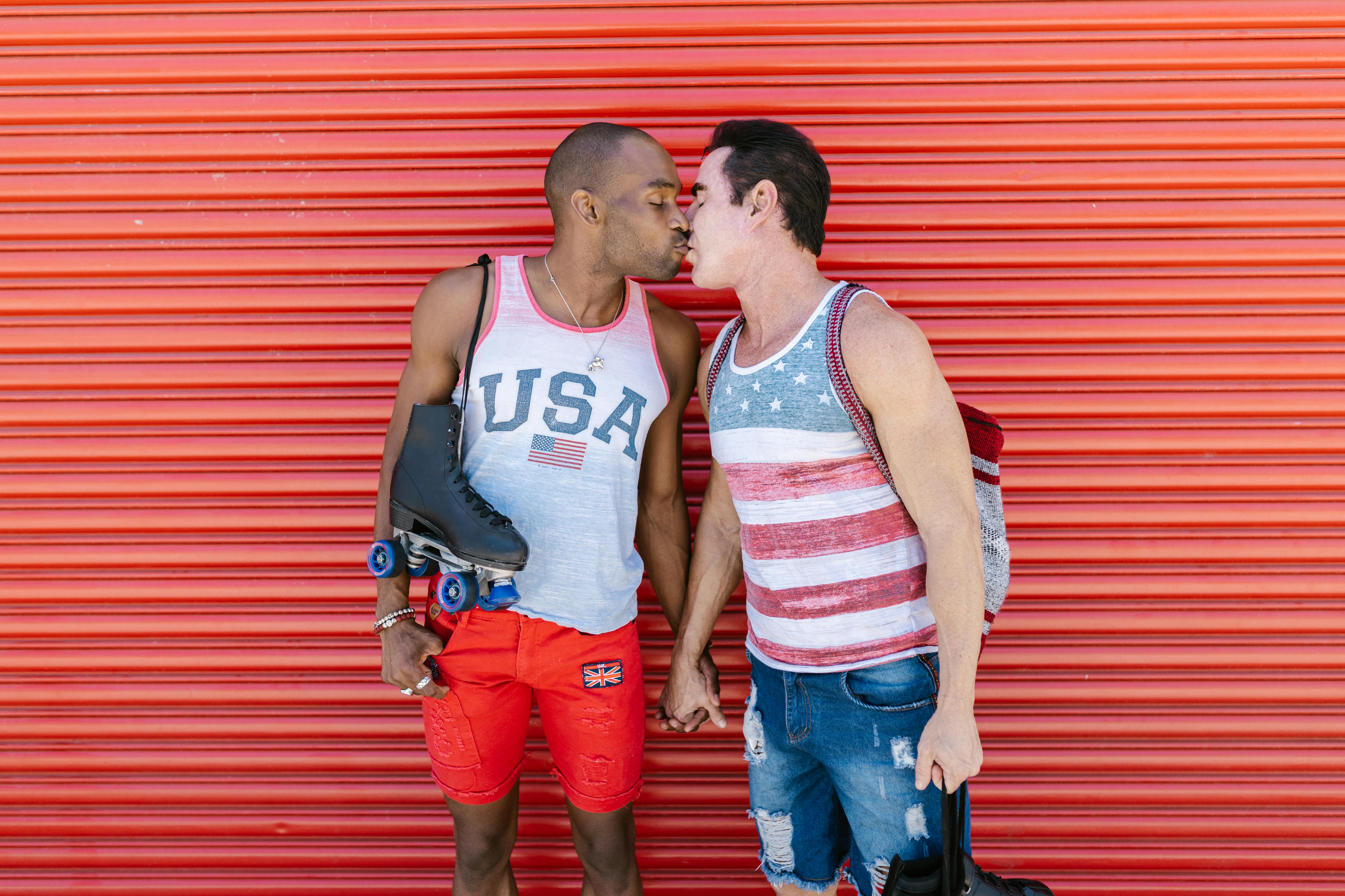 Interracial gay couple sharing a kiss outdoors by a red garage door, showing love and diversity.