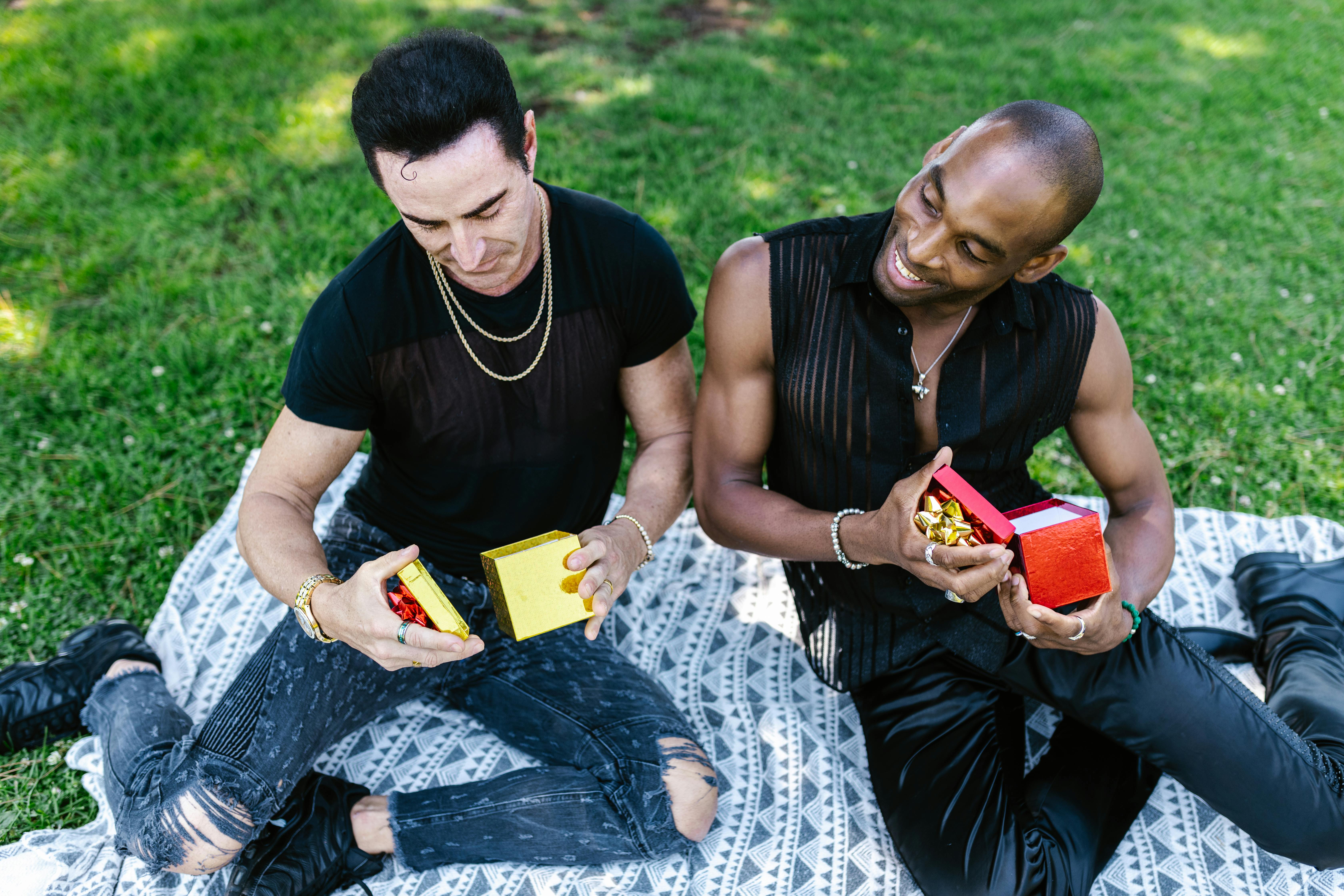 A happy interracial gay couple sharing gifts while sitting on a blanket in a sunny park.