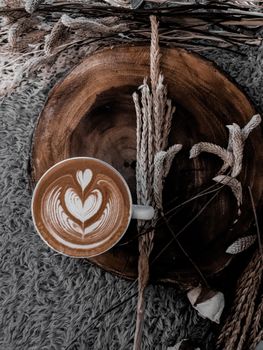 A beautiful latte with intricate latte art surrounded by wheat stalks on a rustic wooden surface.