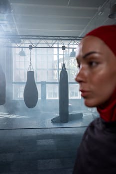 A female boxer wearing a hijab focuses in a misty gym with punching bags.