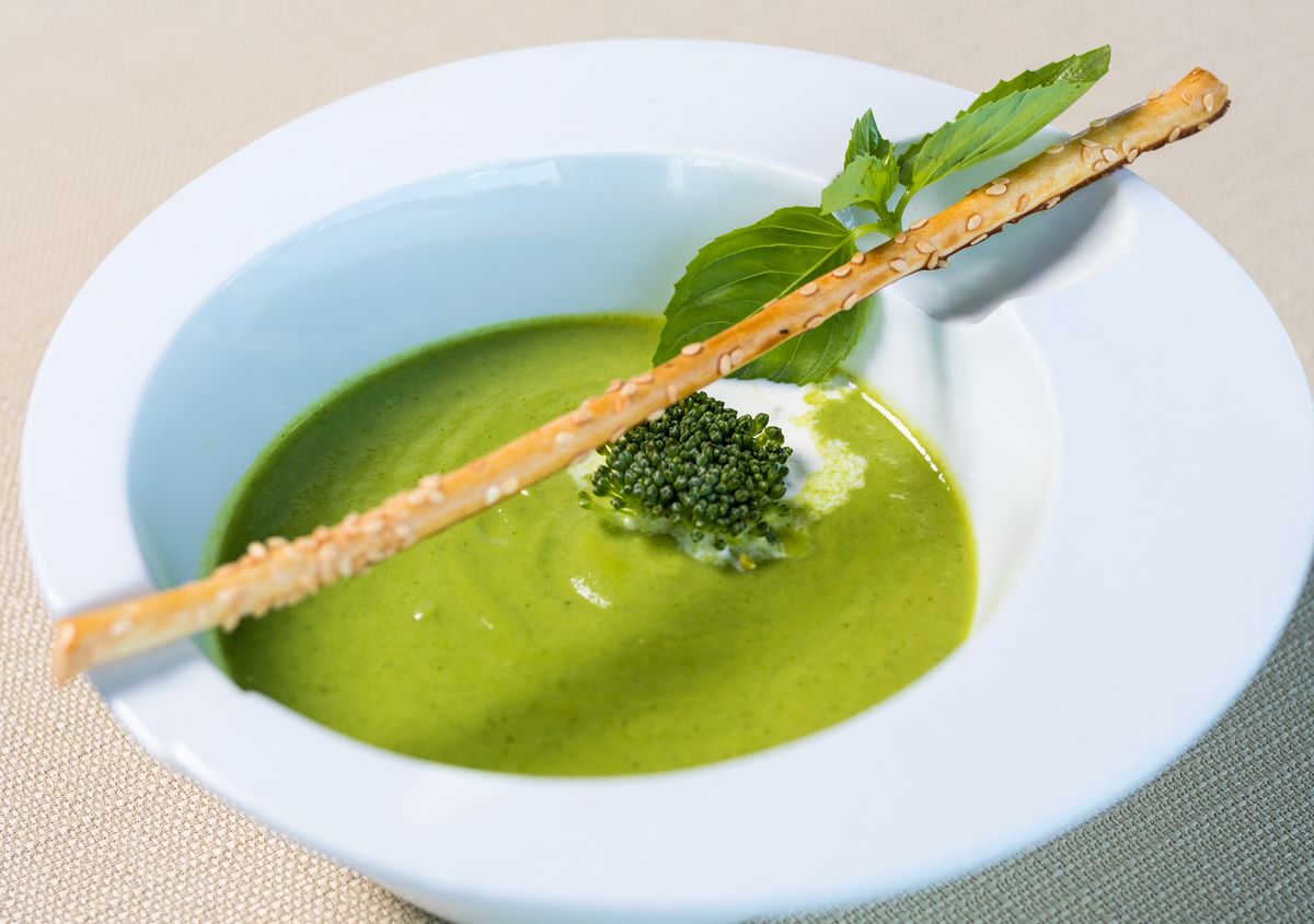 Vivid green broccoli cream soup with a floret, cream dollop, basil and sesame breadstick