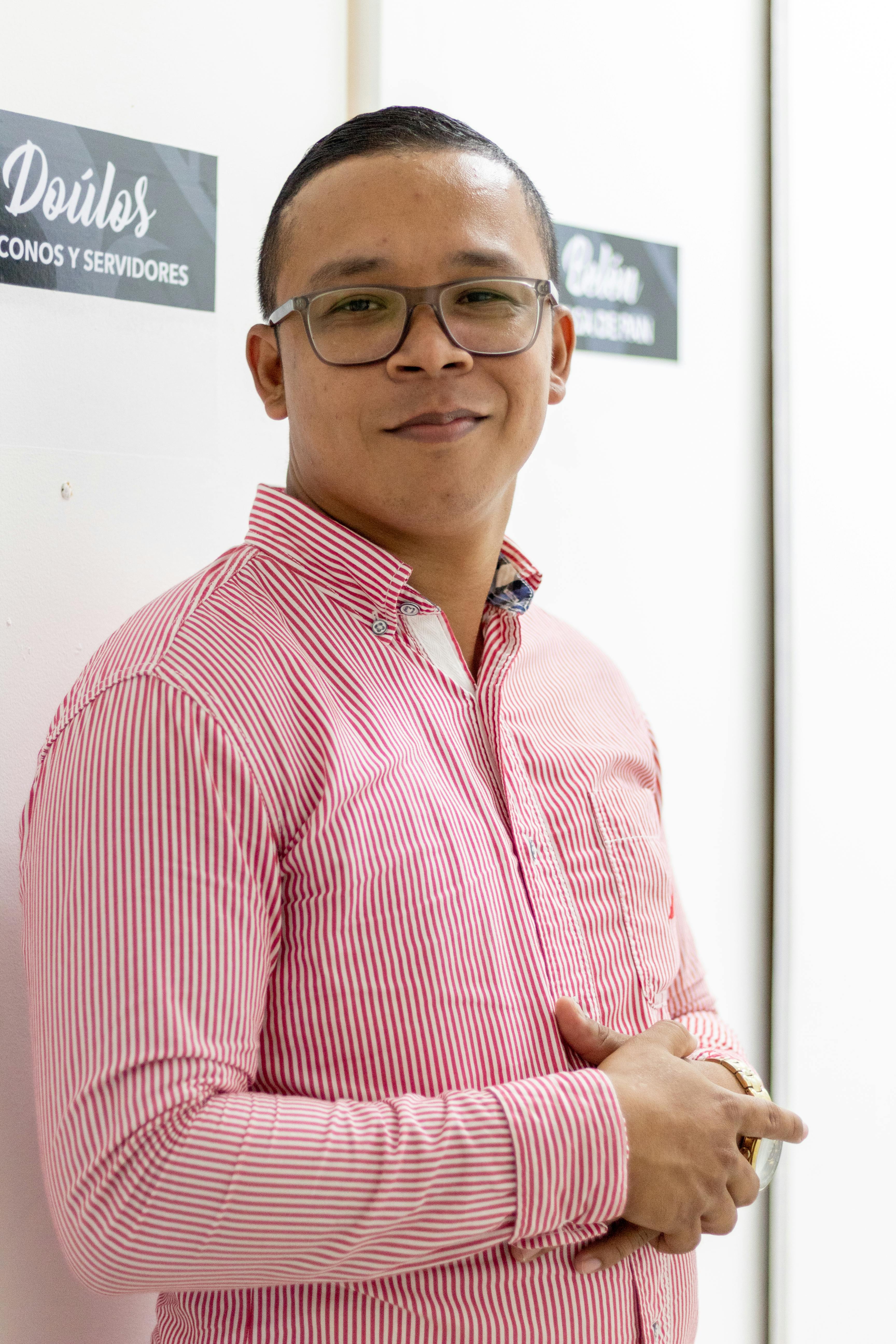 Smiling man in striped shirt and glasses leaning against a wall indoors.