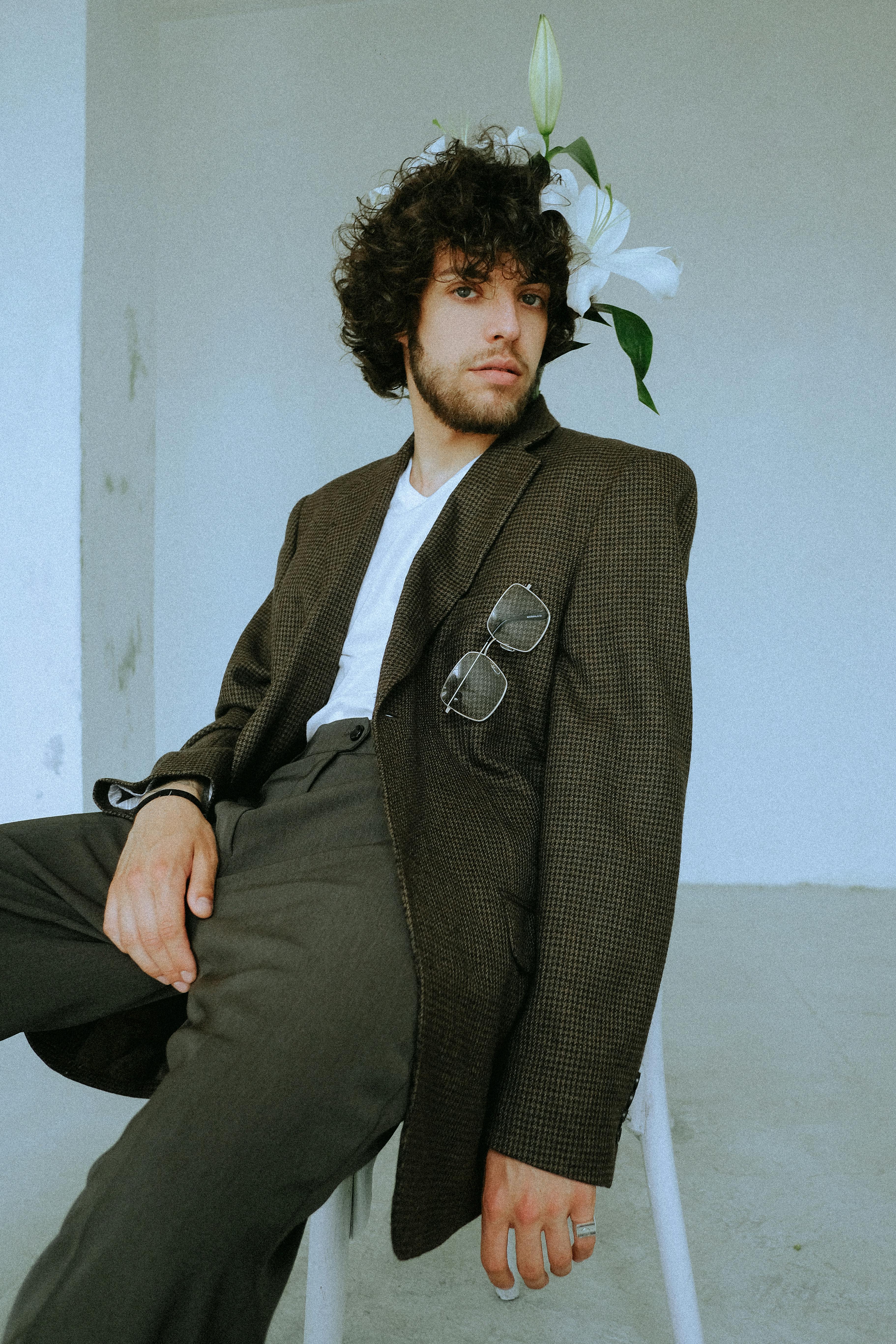 A fashionable man in a suit sits indoors, adorned with lilies for a modern aesthetic.