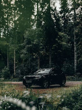 A luxury black car parked on a gravel road surrounded by lush green trees in the woods.