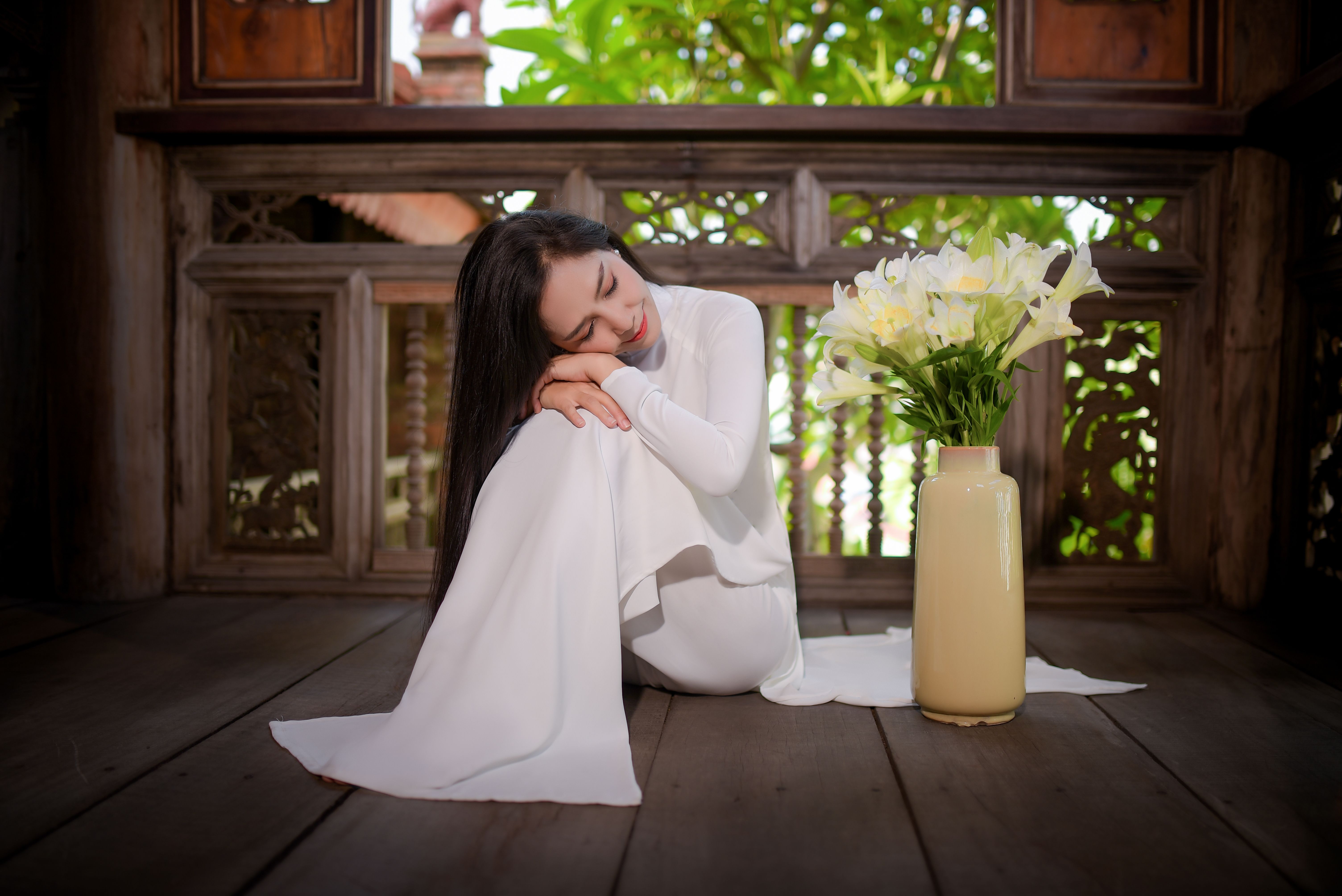 Woman in a white Ao Dai sitting gracefully beside a vase of lilies, exuding tranquility.