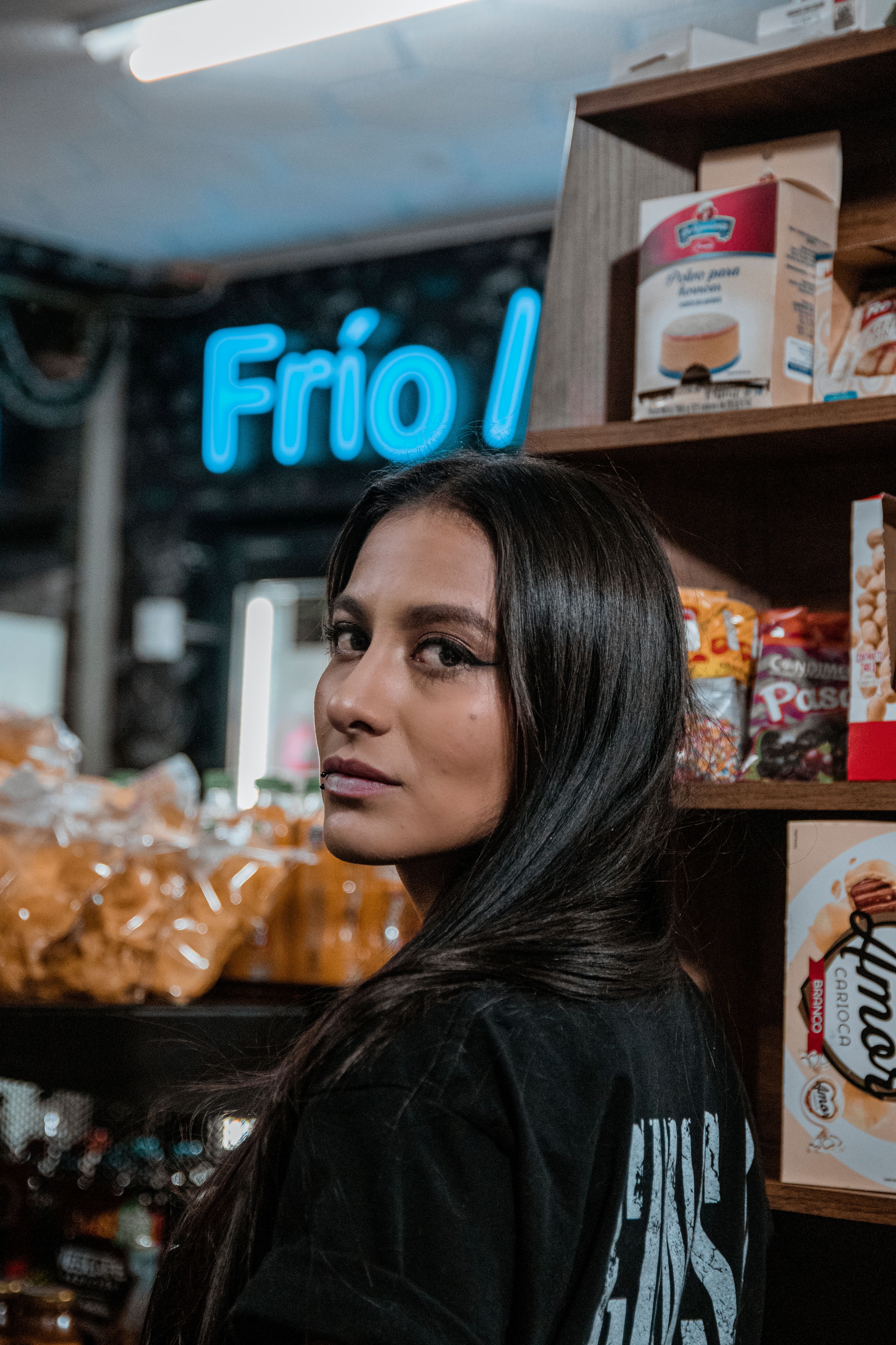 Portrait of a woman in a store with a Frío neon sign and snack shelves.