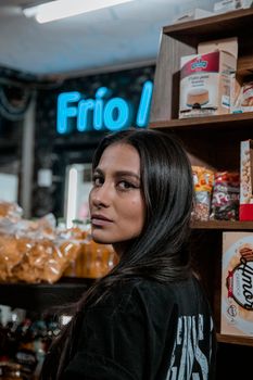 Portrait of a woman in a store with a Frío neon sign and snack shelves.