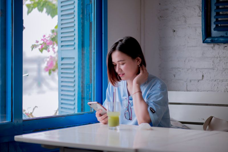 A young woman in a light blue shirt checks her smartphone while enjoying a drink by a window in a quaint cafe.