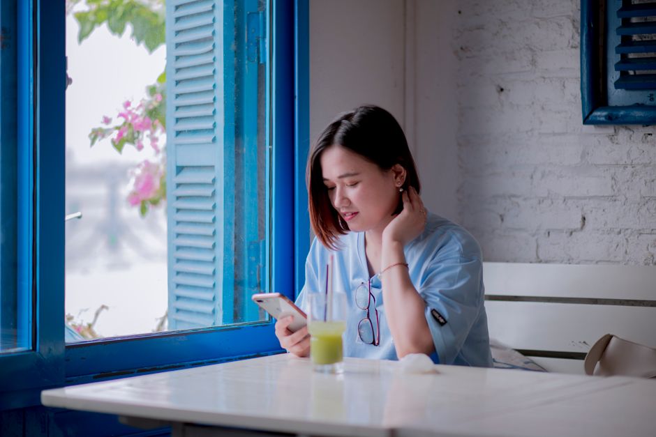 A young woman in a light blue shirt checks her smartphone while enjoying a drink by a window in a quaint cafe. A young woman in a light blue shirt checks her smartphone while enjoying a drink by a window in a quaint cafe.