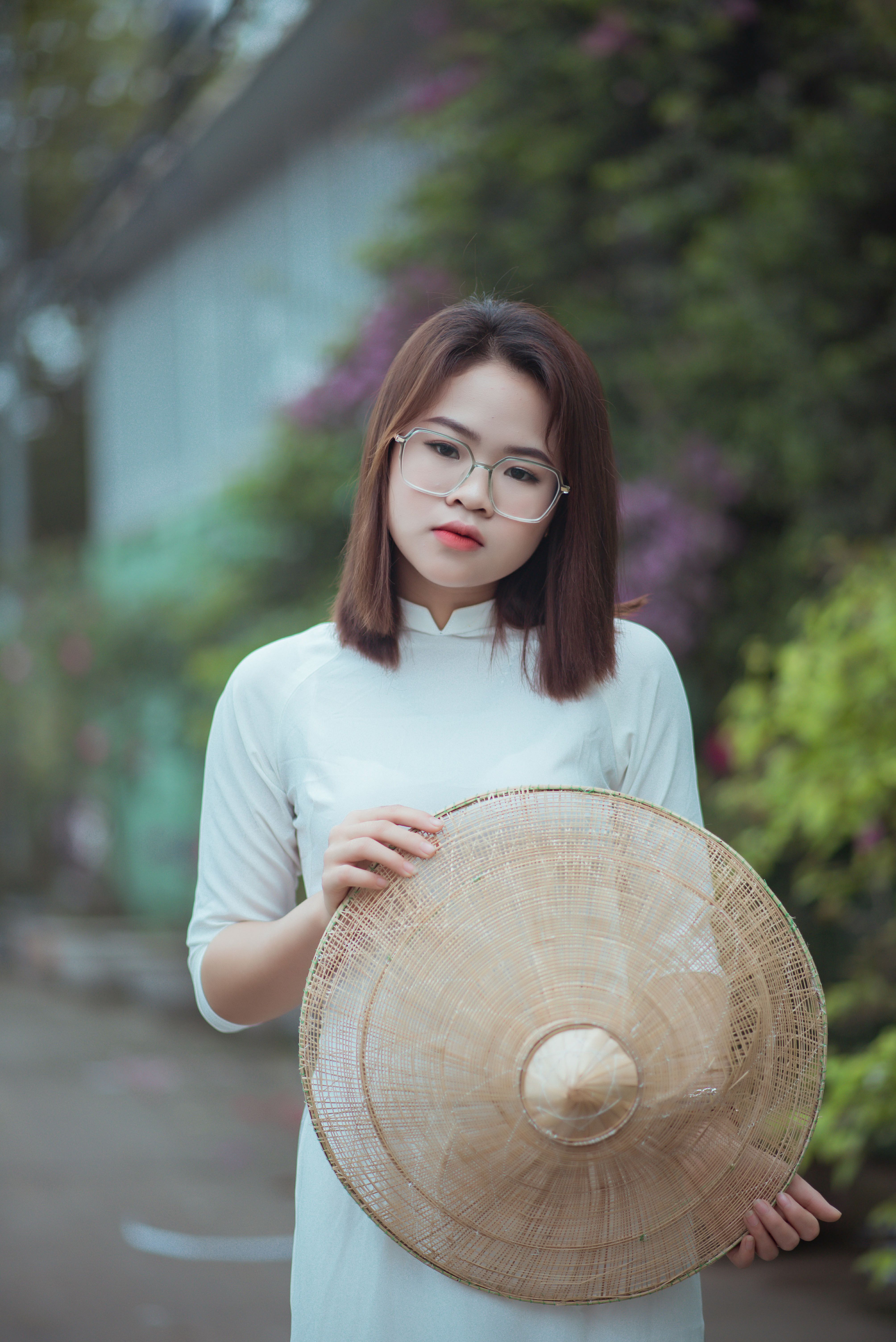 A young Asian woman in a white dress and glasses holding a conical hat outdoors.