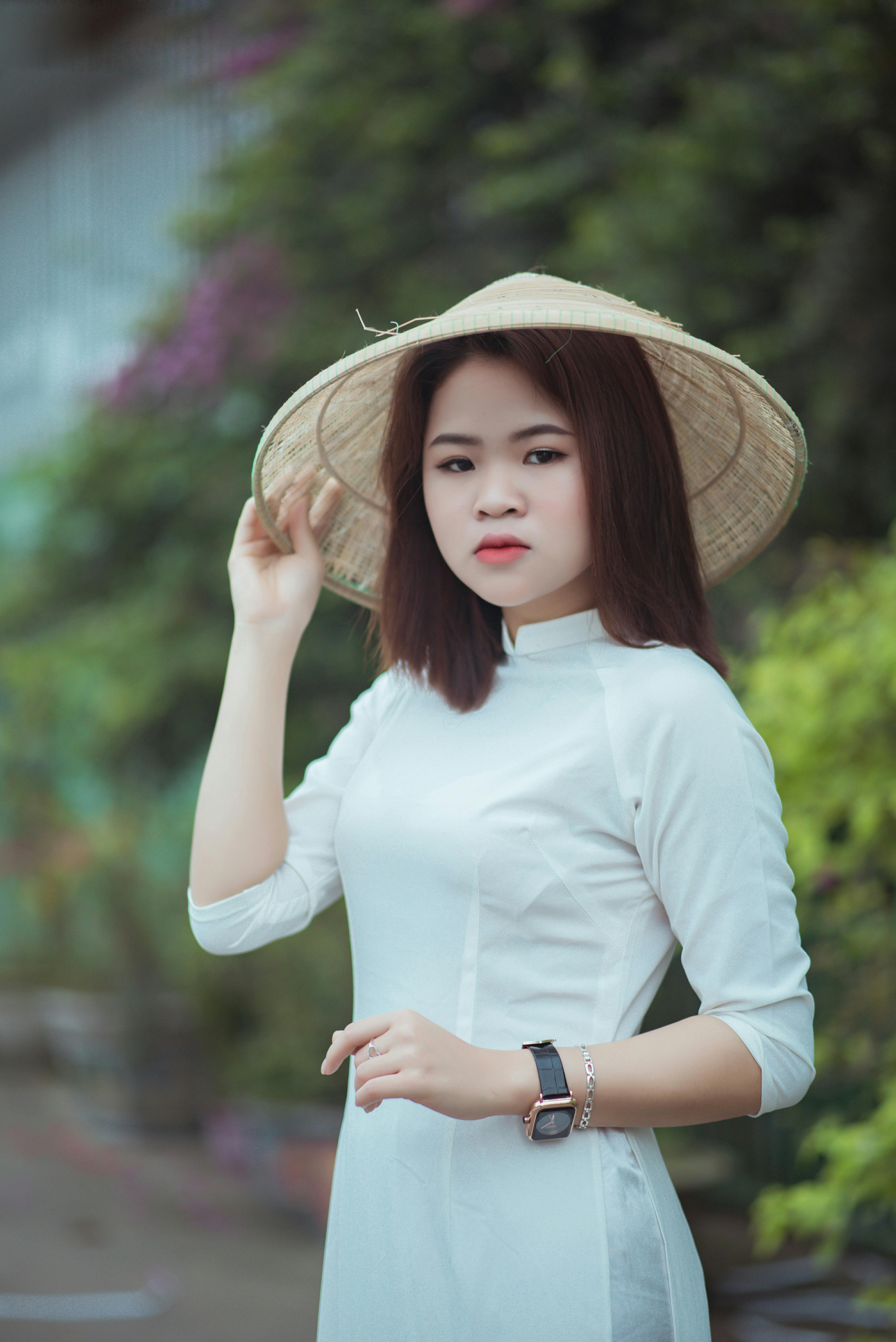 Portrait of an Asian woman in a white dress and hat, captured outdoors with a natural backdrop.