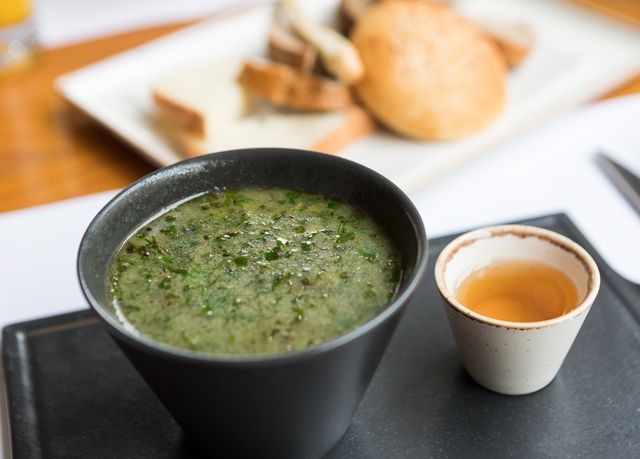 A bowl of herbal green soup served with bread and a side dip, offering a delicious meal.